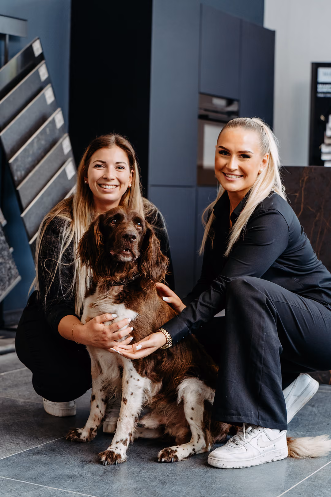 Zwei lächelnde Frauen in schwarzer Kleidung knien auf dem Boden und halten einen braun-weißen Hund in einem modernen Raum mit dunklen Wänden und Steinfliesen.