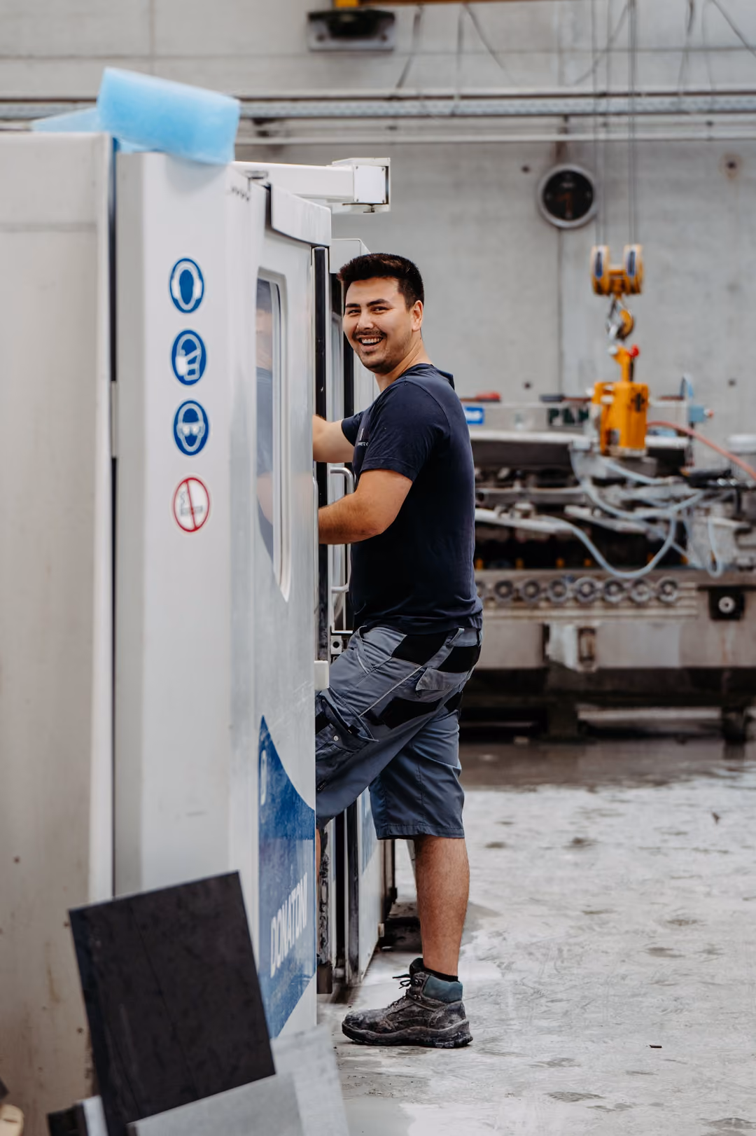 Mitarbeiter in Arbeitskleidung bedient eine Maschine in einer Fabrikhalle und lächelt zur Kamera.