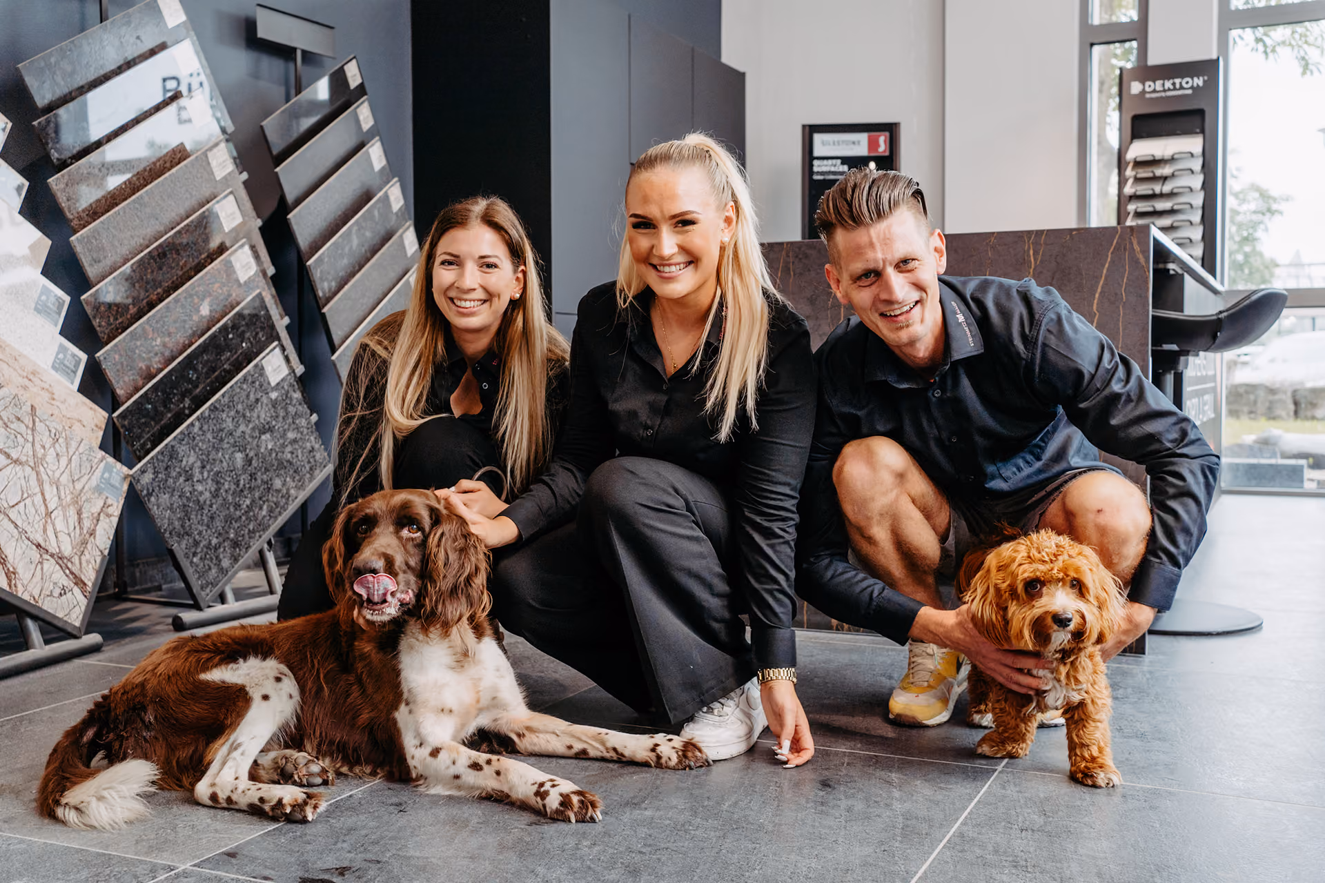 Drei lächelnde Personen in schwarzer Kleidung hocken auf grauem Boden, jede mit einem Hund: ein großer braun-weißer Hund liegt links, ein kleiner brauner Hund wird rechts gehalten.