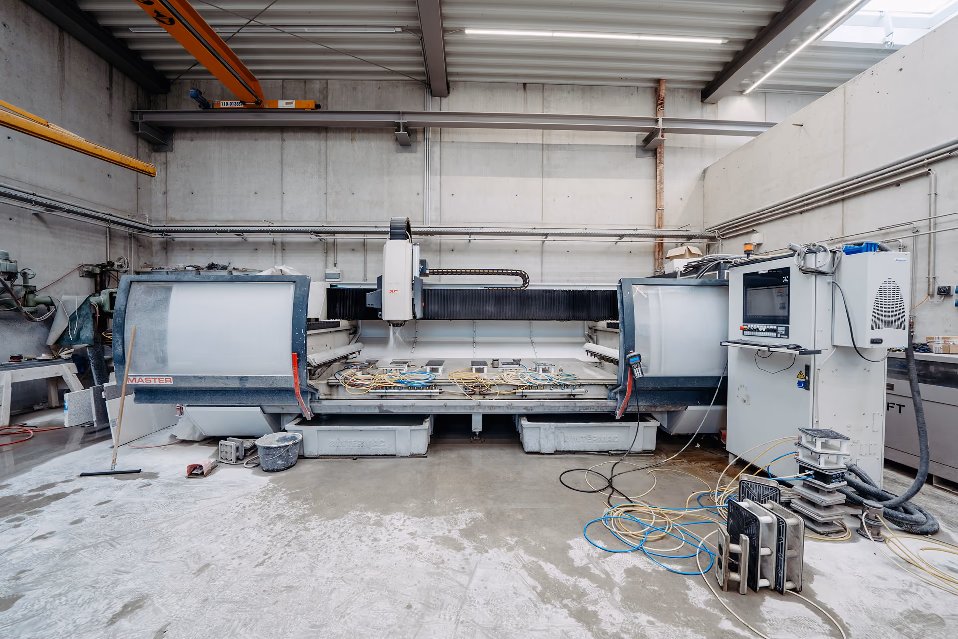 Industrielle Maschine mit Computersteuerung in einer Werkhalle mit Betonboden und sichtbarer Elektronik und Kabeln an der Seite.