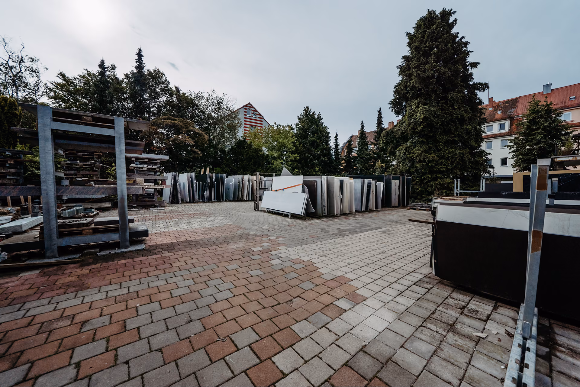 Außenlager eines Unternehmens mit gestapelten großen Steinplatten und Baumaterialien auf gepflastertem Boden, umgeben von Bäumen und Gebäuden.