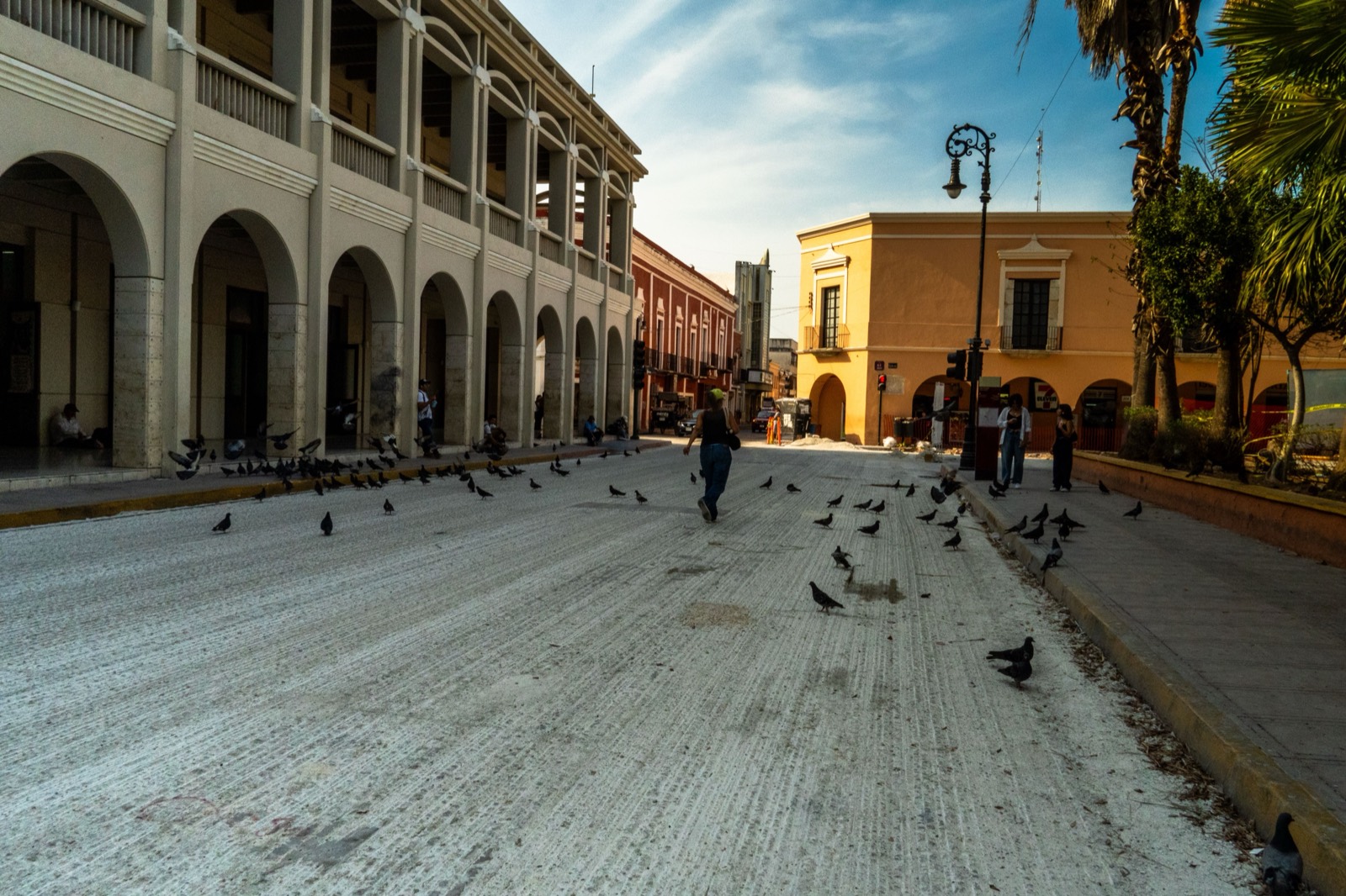 People walking across a colonial plaza in Mérida, Yucatán, pigeons on the pavement and arcaded civic buildings behind