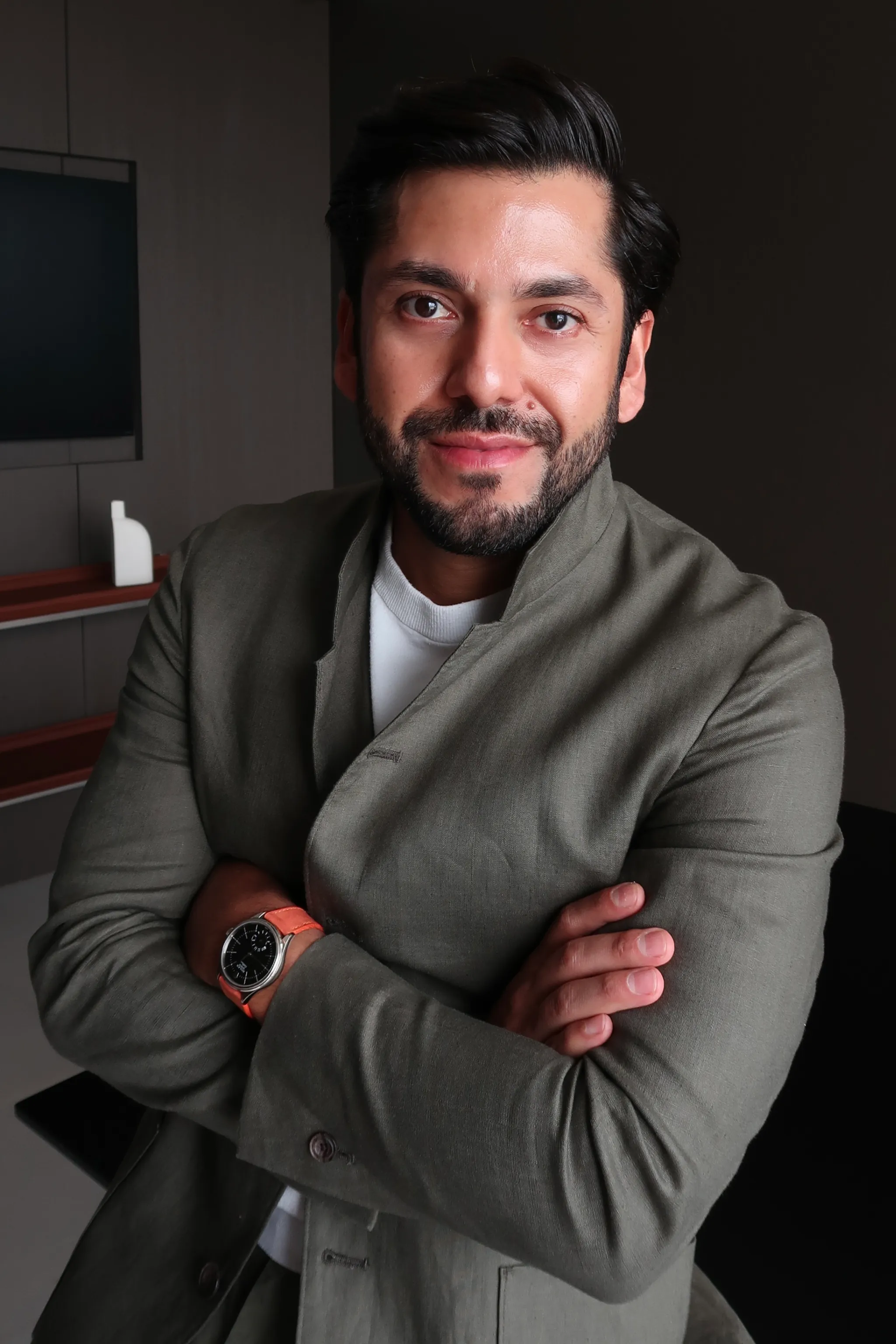 Man with dark hair and beard wearing a light gray jacket with arms crossed, smiling at camera.