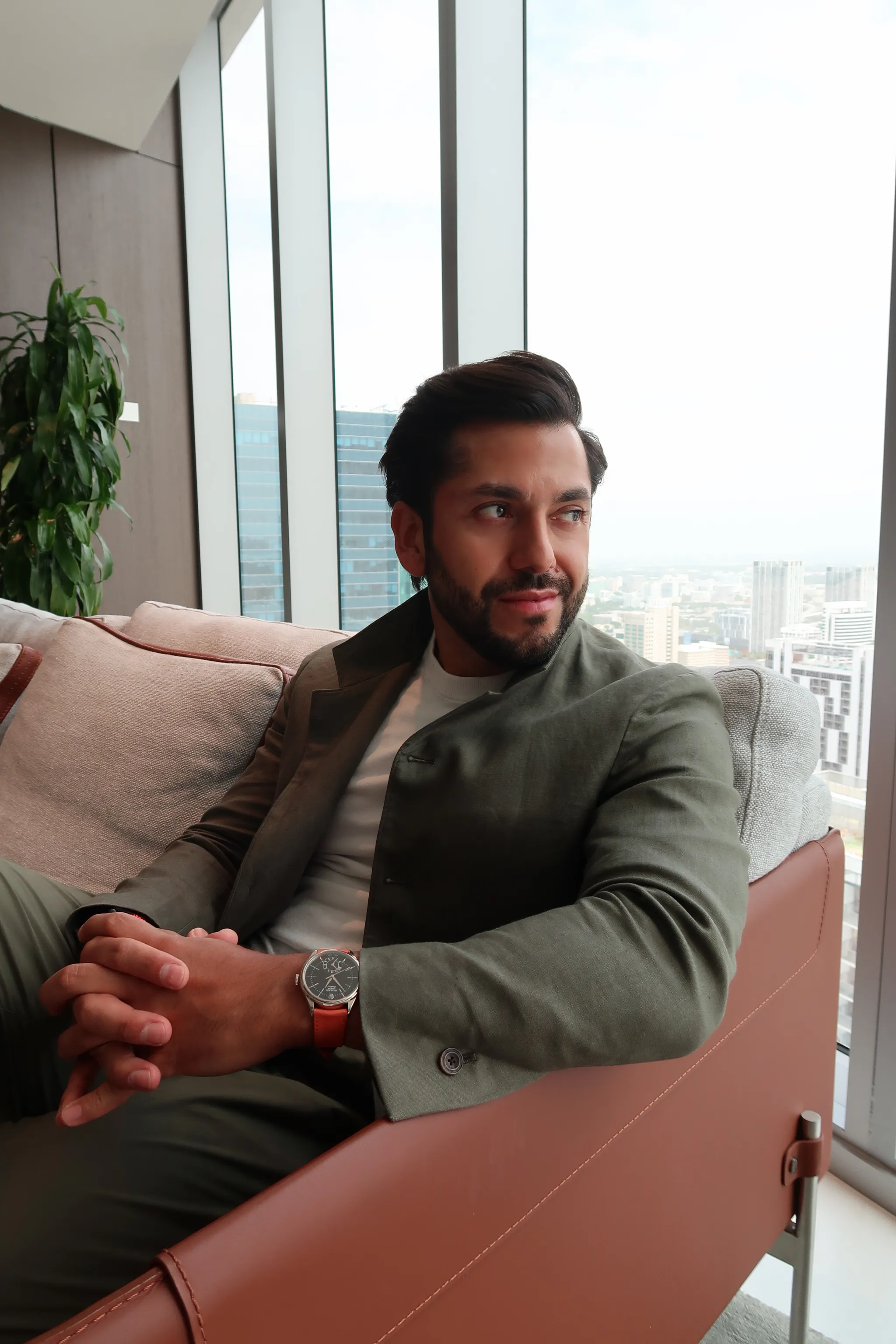 Man with dark hair and beard sitting on a couch near a large window overlooking a cityscape.