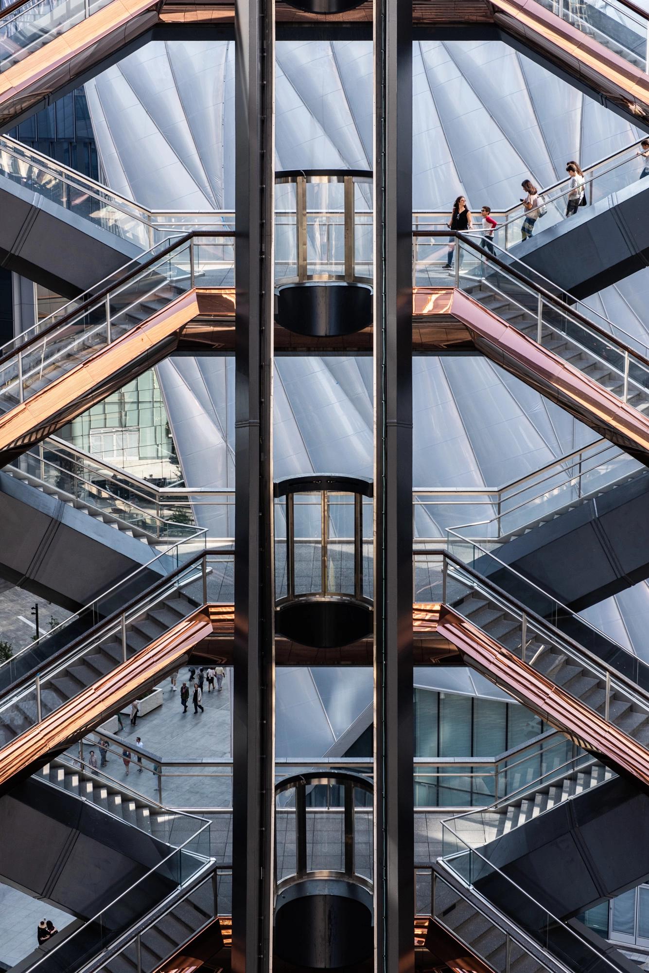 Symmetrical modern staircases with glass elevators and copper-colored railings in an urban architectural setting.