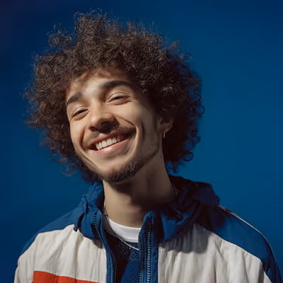 Young man with curly hair and a goatee smiling against a dark blue background.