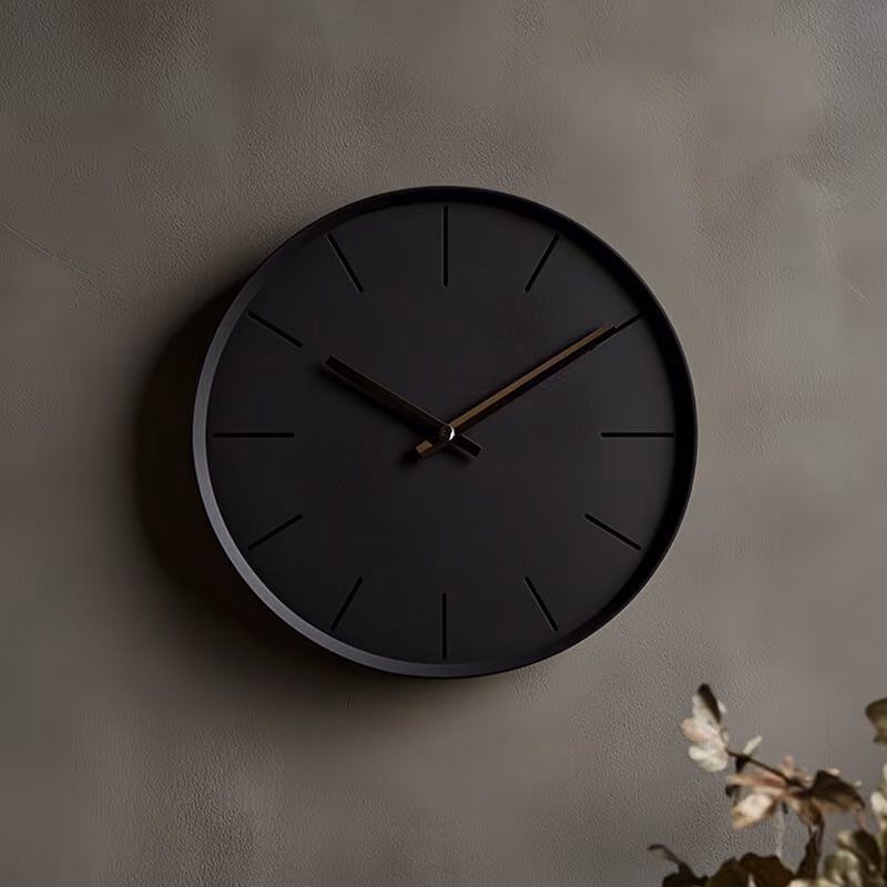 Minimalist black wall clock with no numbers on a gray textured wall near dried leaves.