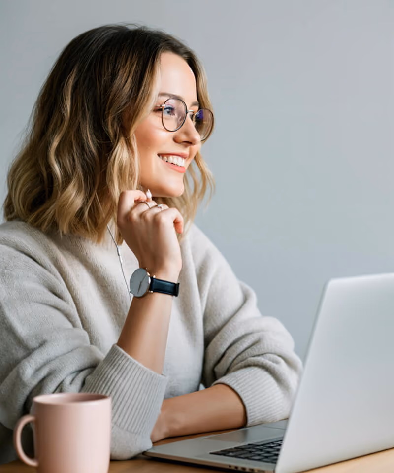 Smiling woman with glasses wearing earbuds, using a laptop at a desk with a pink mug.