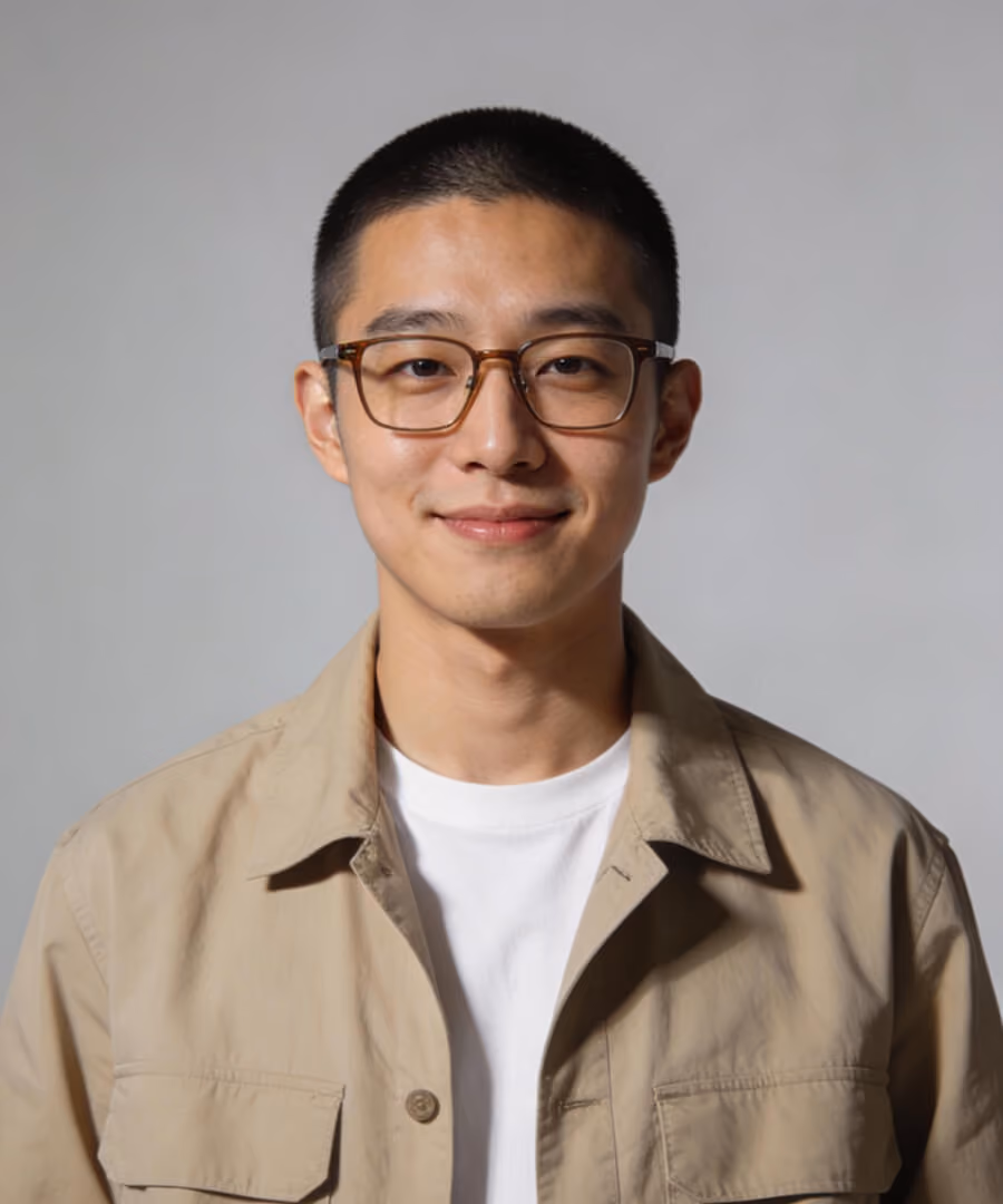 Smiling young man with short hair and glasses wearing beige jacket and white shirt.