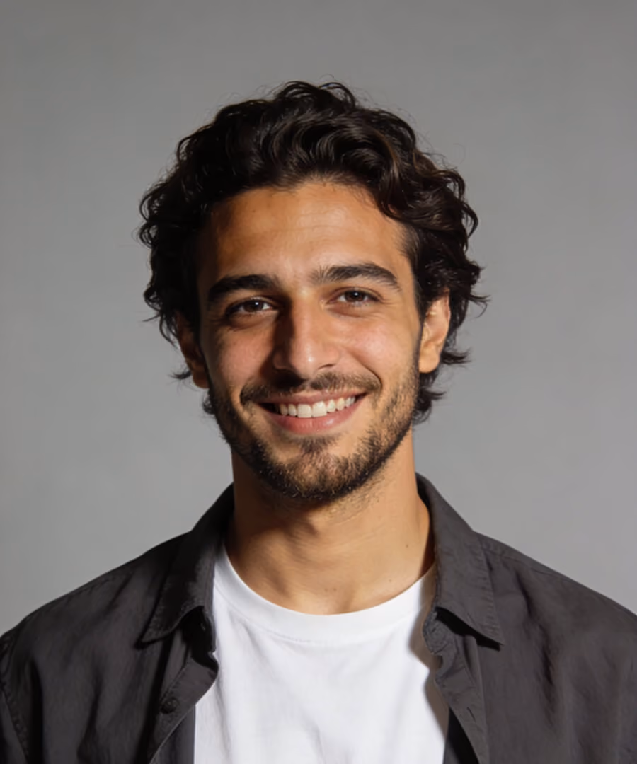 Smiling man with curly dark hair, beard, white shirt, and dark open jacket on gray background.