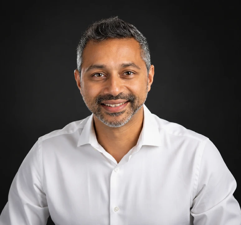 Smiling man with salt-and-pepper hair and beard wearing a white button-up shirt against black background.