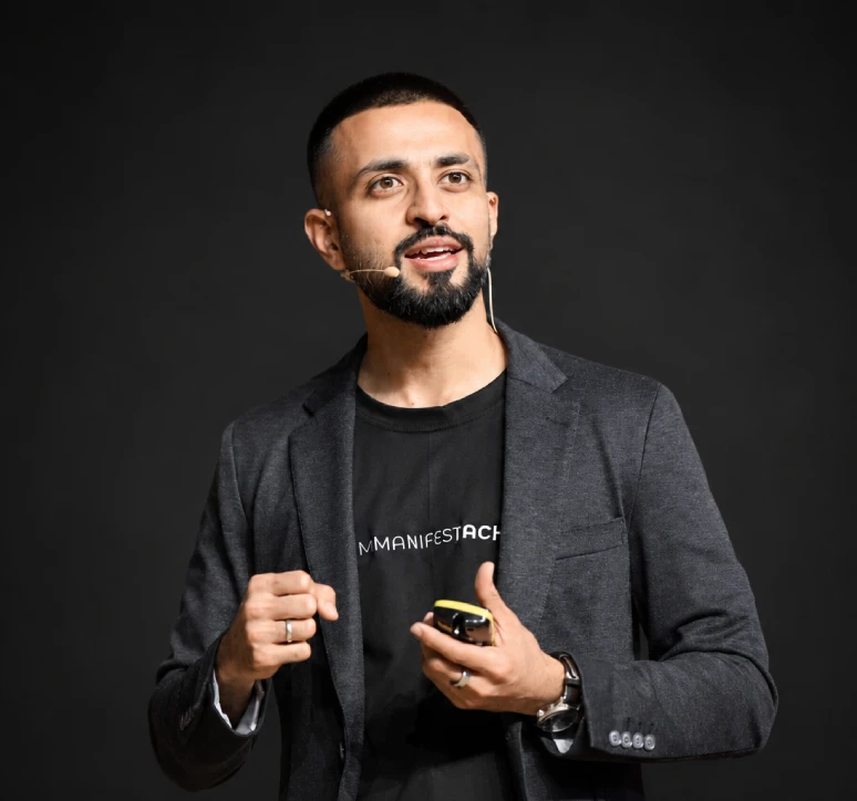 Man with a beard and microphone headset speaking and gesturing during a presentation against black background.