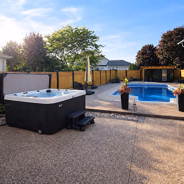 Outdoor patio with a hot tub on the left and a blue swimming pool surrounded by a wooden fence.