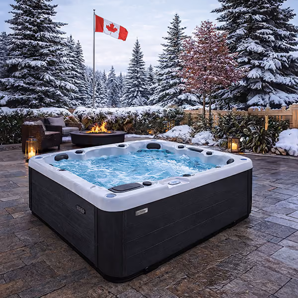 Outdoor hot tub bubbling on stone patio with fire pit, chairs, snowy pine trees, and Canadian flag in background.