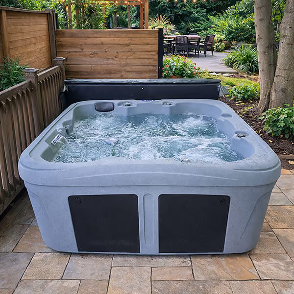 Outdoor gray hot tub with bubbling water on a tiled patio surrounded by wooden fence and greenery.