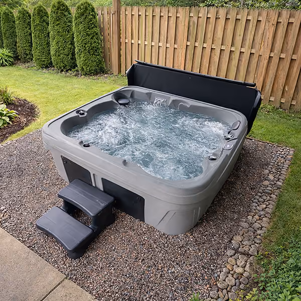 Outdoor gray hot tub with bubbling water and black steps, set on gravel in a fenced backyard.