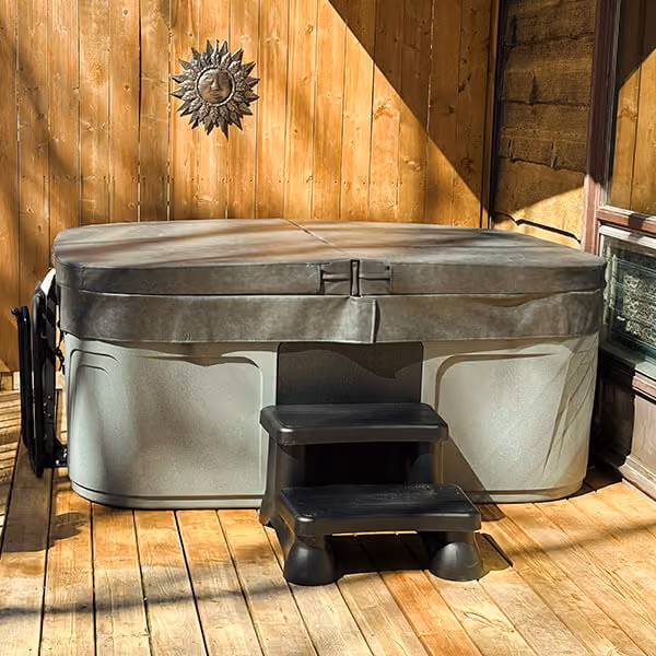 Gray hot tub with closed cover and black steps on wooden deck against wood panel wall.