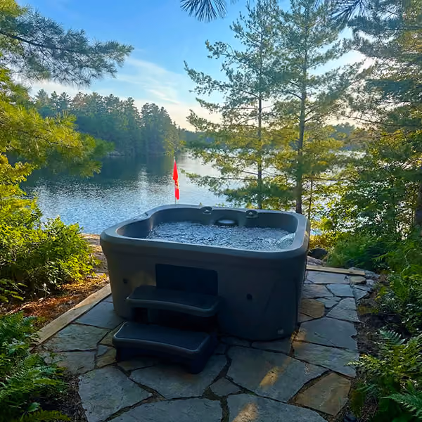 Outdoor hot tub with bubbling water on stone patio by a lake surrounded by trees under blue sky.
