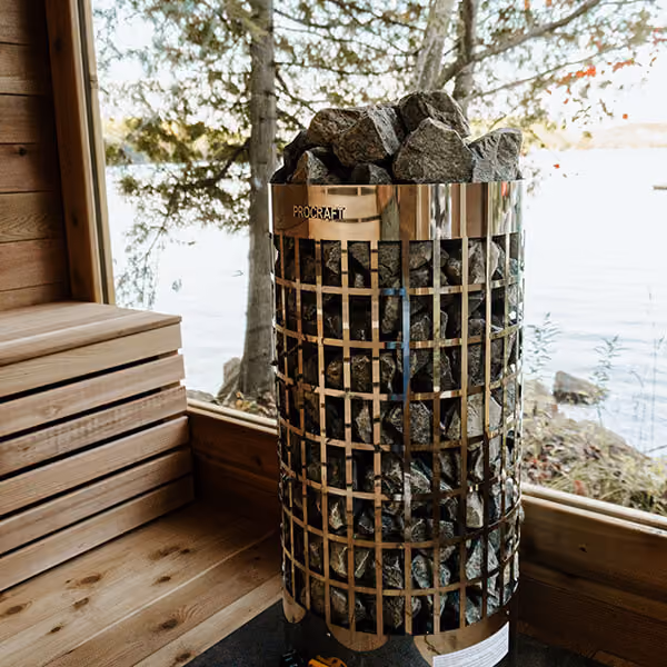 Modern sauna heater filled with sauna stones inside a wooden sauna room with a window view of trees and water.