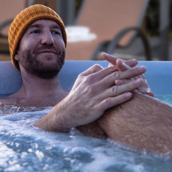 Man with a brown beanie relaxing with arms around knees in bubbling water outdoors.