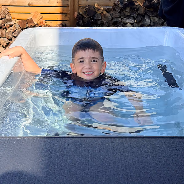 Young boy smiling and relaxing in a small outdoor water tub with wood stacked in the background.