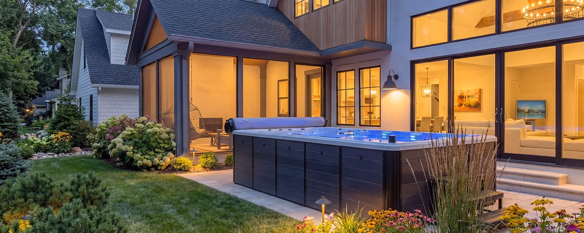 Modern backyard with a large swim spa next to a house lit warmly with large windows.