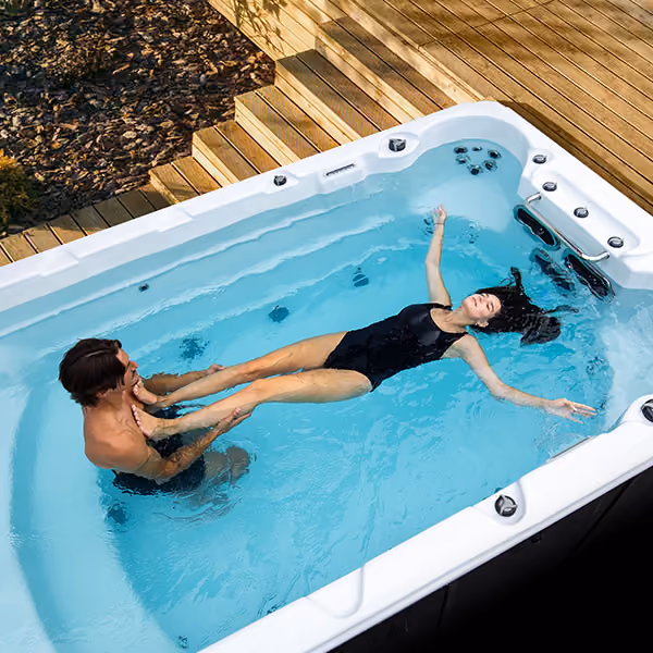 Man and woman in black swimsuits enjoying water exercise in a rectangular outdoor swim spa.