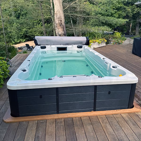 Rectangular outdoor swim spa with clear water on wooden deck surrounded by trees and plants.