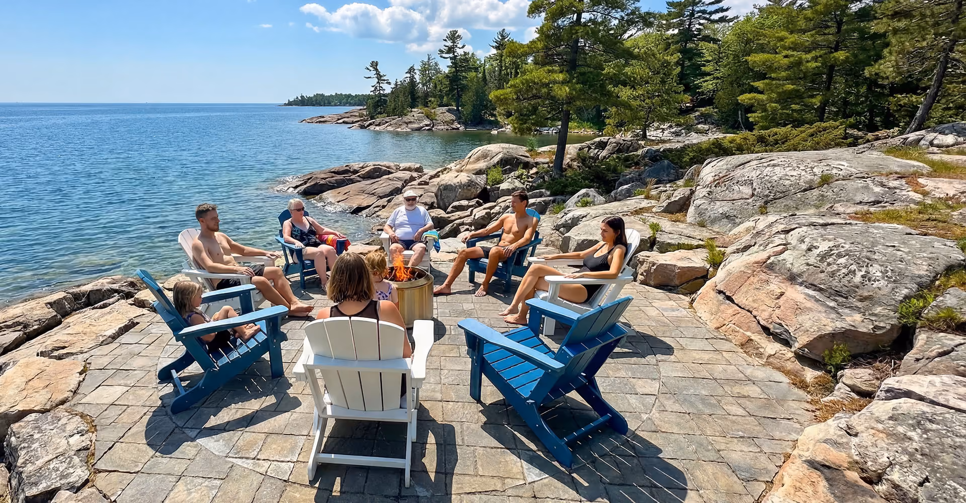 Group of people sitting in Adirondack chairs around a fire pit by lake shore with rocks and trees.