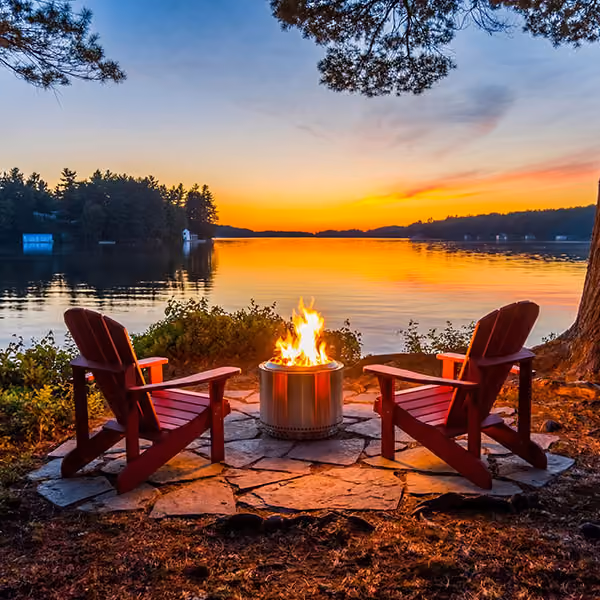 Two wooden chairs around a fire pit on a lakeside at sunset with trees and calm water.