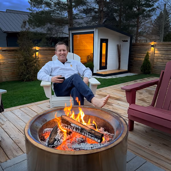 Man sitting barefoot in white chair by a fire pit on wooden deck in backyard at dusk.