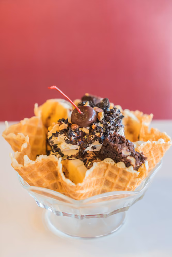 Ice cream sundae in a waffle bowl topped with banana slices, whipped cream, cherry, nuts, and brownie pieces.