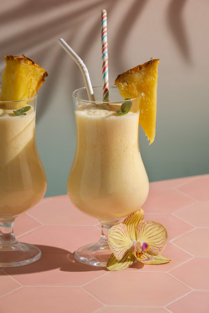 Two creamy pineapple smoothies with pineapple slices, mint leaves, and straws on a pink table.
