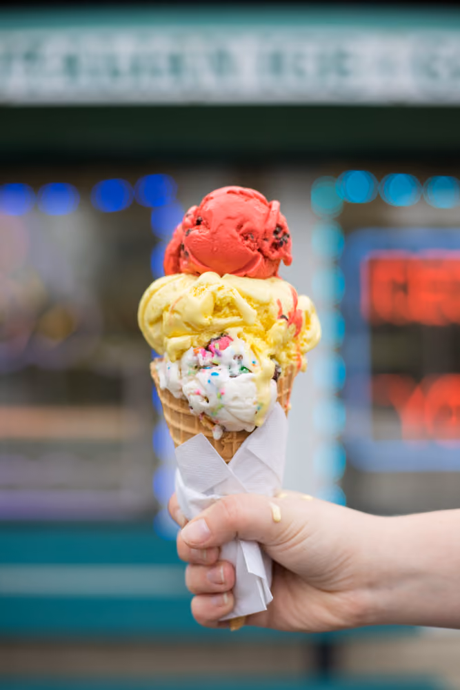 Hand holding a triple scoop ice cream cone with red, yellow, and white scoops in a waffle cone.