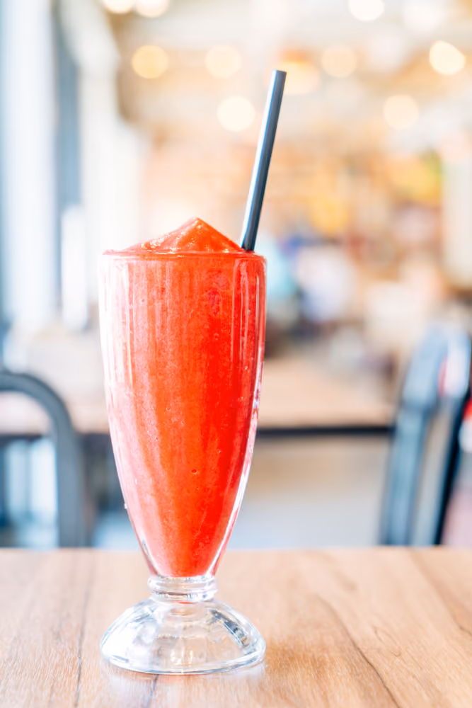 Tall glass of red slushy drink with black straw on wooden table in bright cafe.