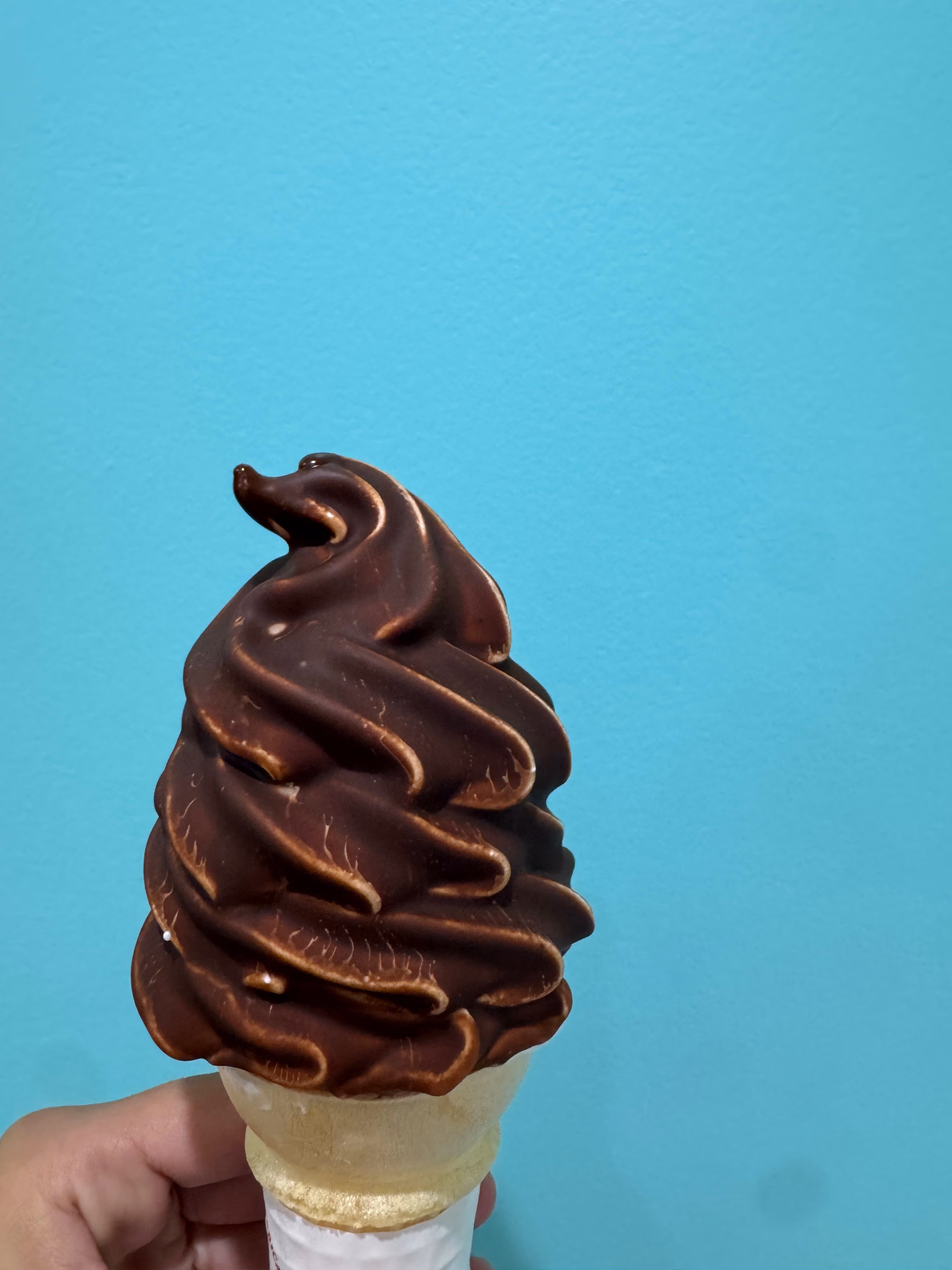 Hand holding a chocolate-dipped soft serve ice cream cone against a blue background.