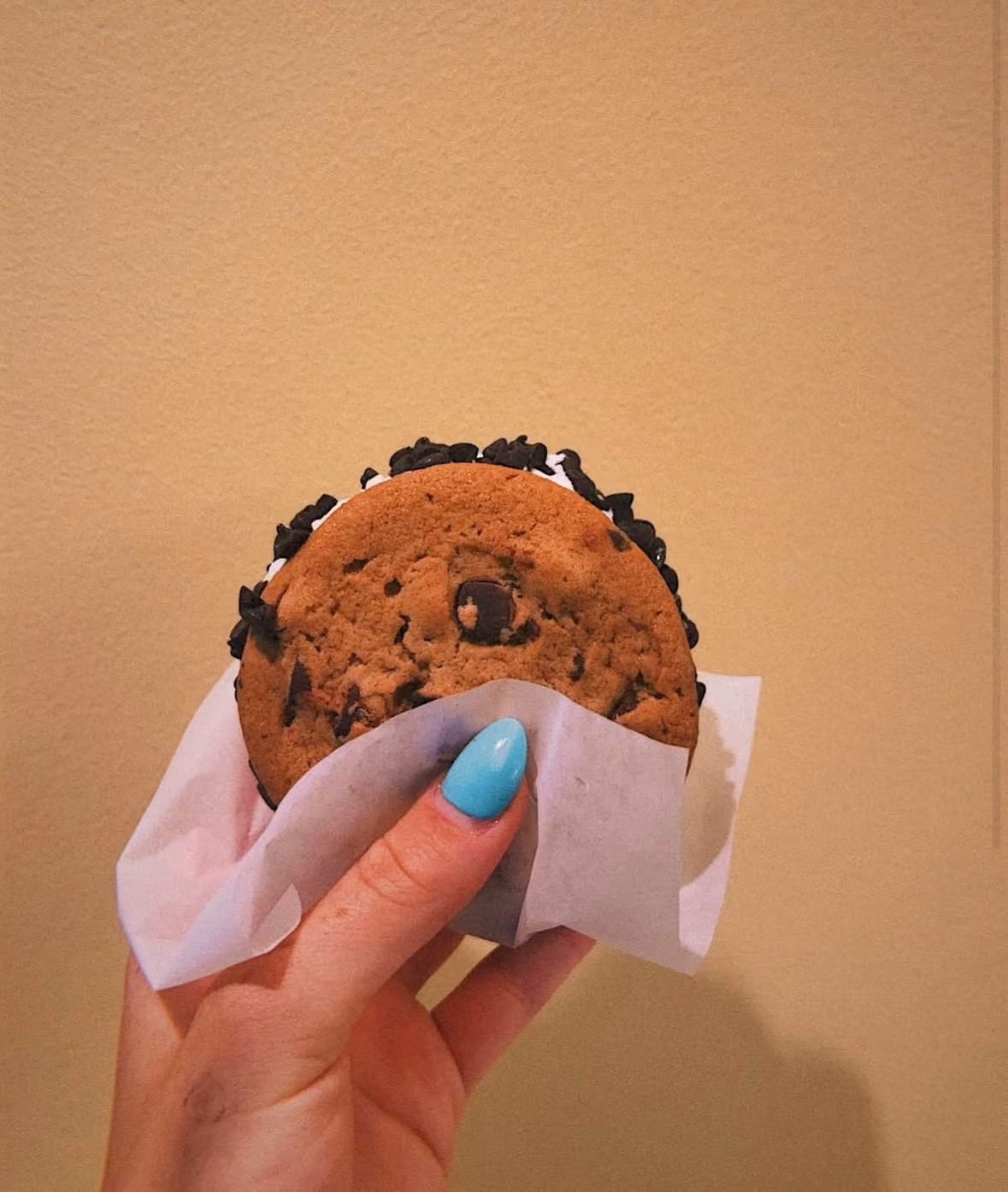 Hand with blue nail polish holding a chocolate chip ice cream sandwich wrapped in paper.