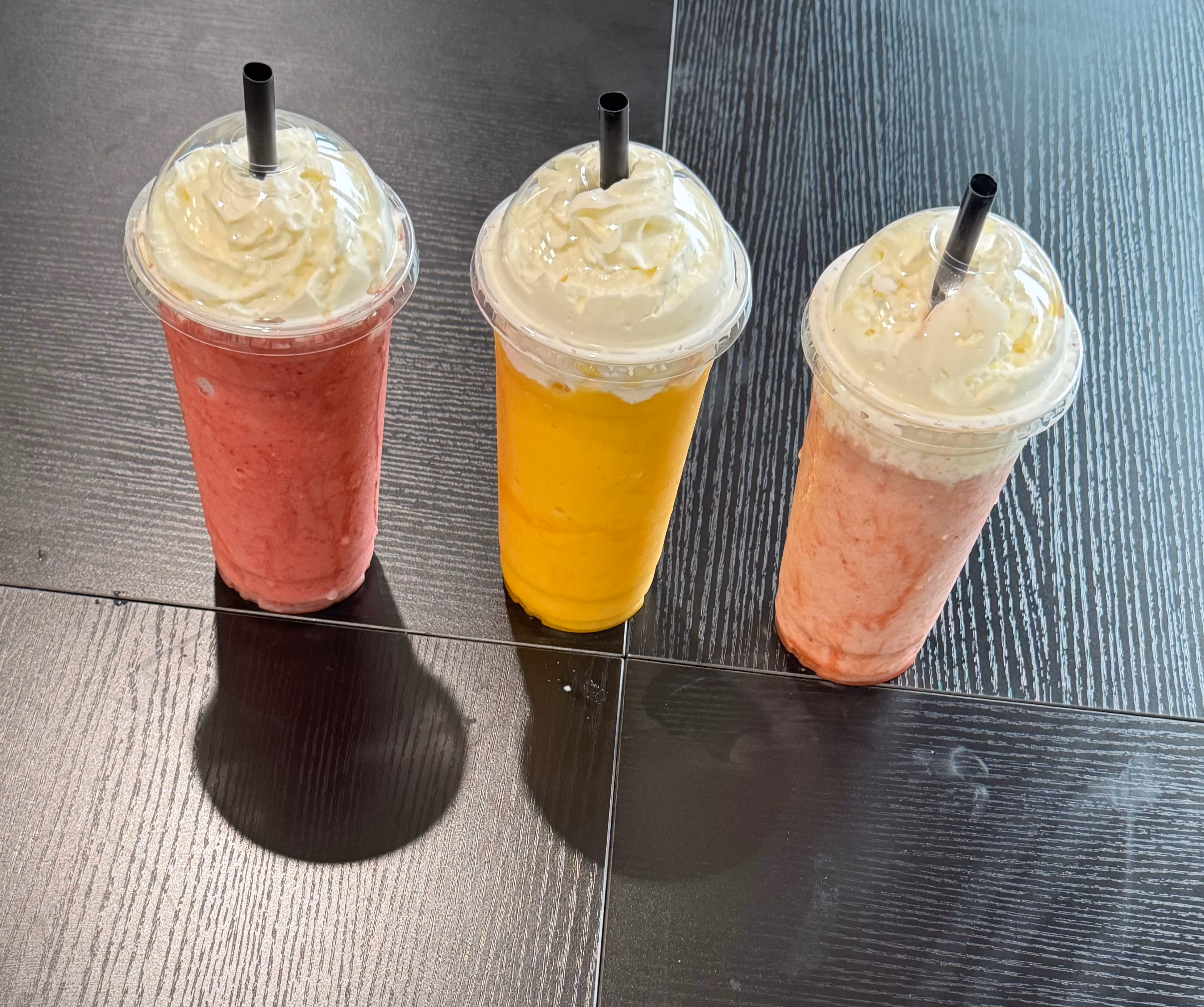 Three smoothie cups with whipped cream and straws on a dark wooden table in red, yellow, and pink colors.