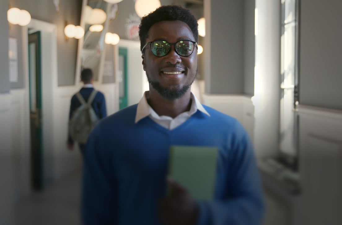 Smiling man wearing glasses and blue sweater holding a book in a hallway with another person blurred behind.