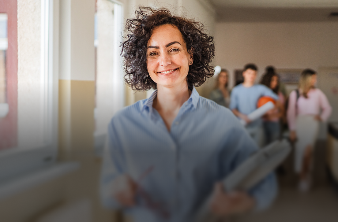Smiling woman with curly hair holding documents, people blurred in background in a hallway.