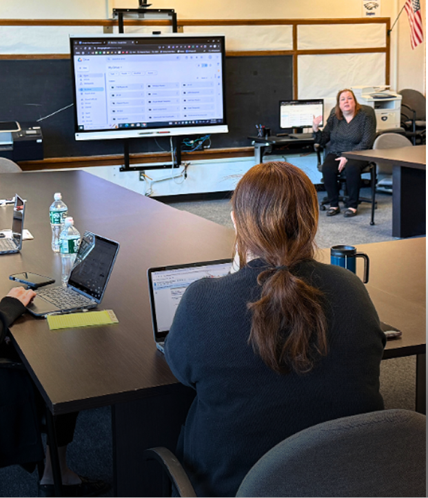 Woman presenting in a classroom with two people seated at a table using laptops and a large screen showing a user interface.