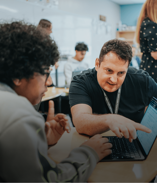 Teacher pointing at laptop screen while helping student in classroom with other students in background.