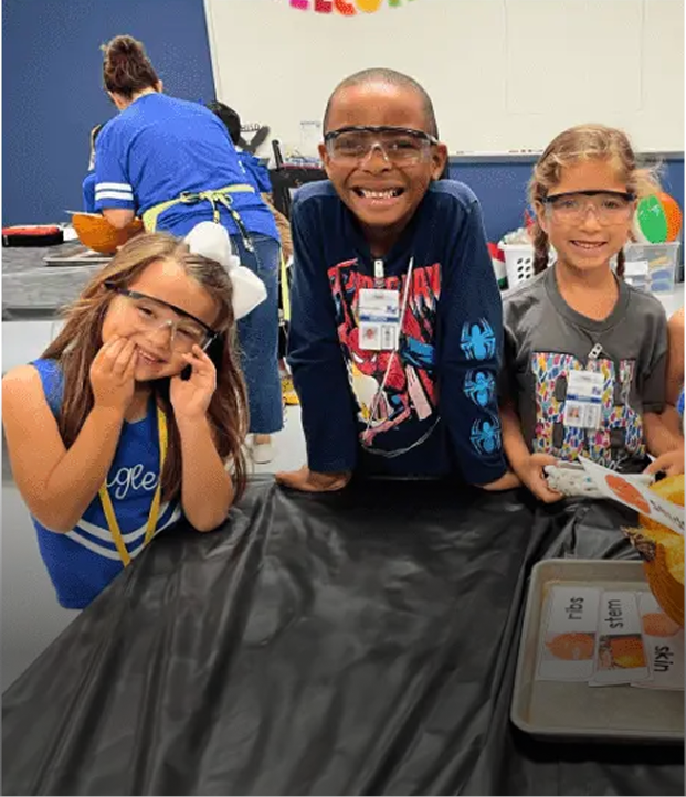 Three children wearing safety goggles smiling and leaning on a table in a classroom.