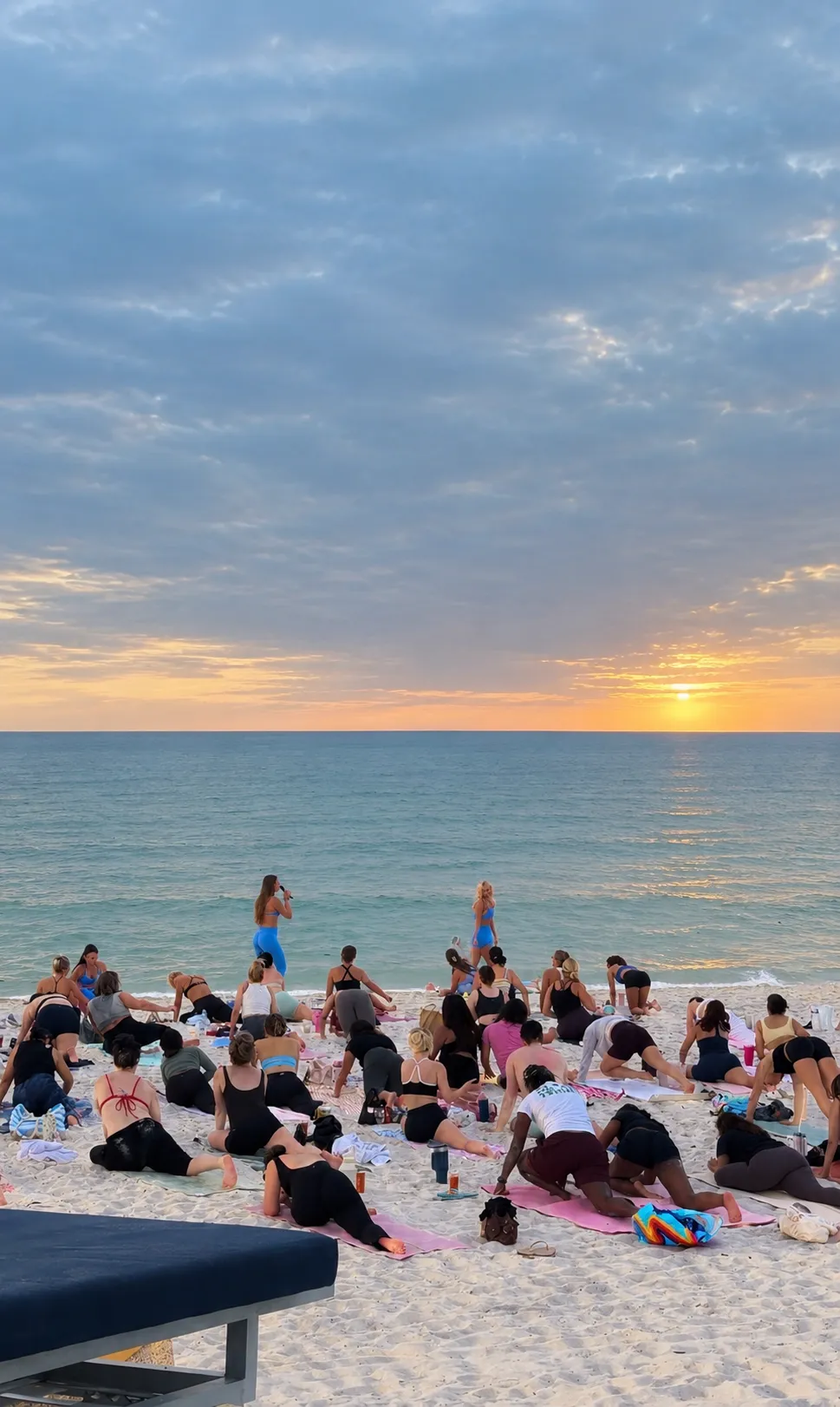 A Sculpted beach class on mats at sunset, two instructors cueing from the shoreline