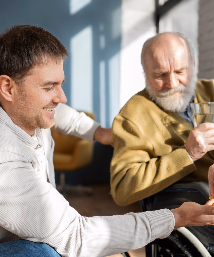 Younger man smiling and holding the hand of an elderly man in a wheelchair who is holding a glass.