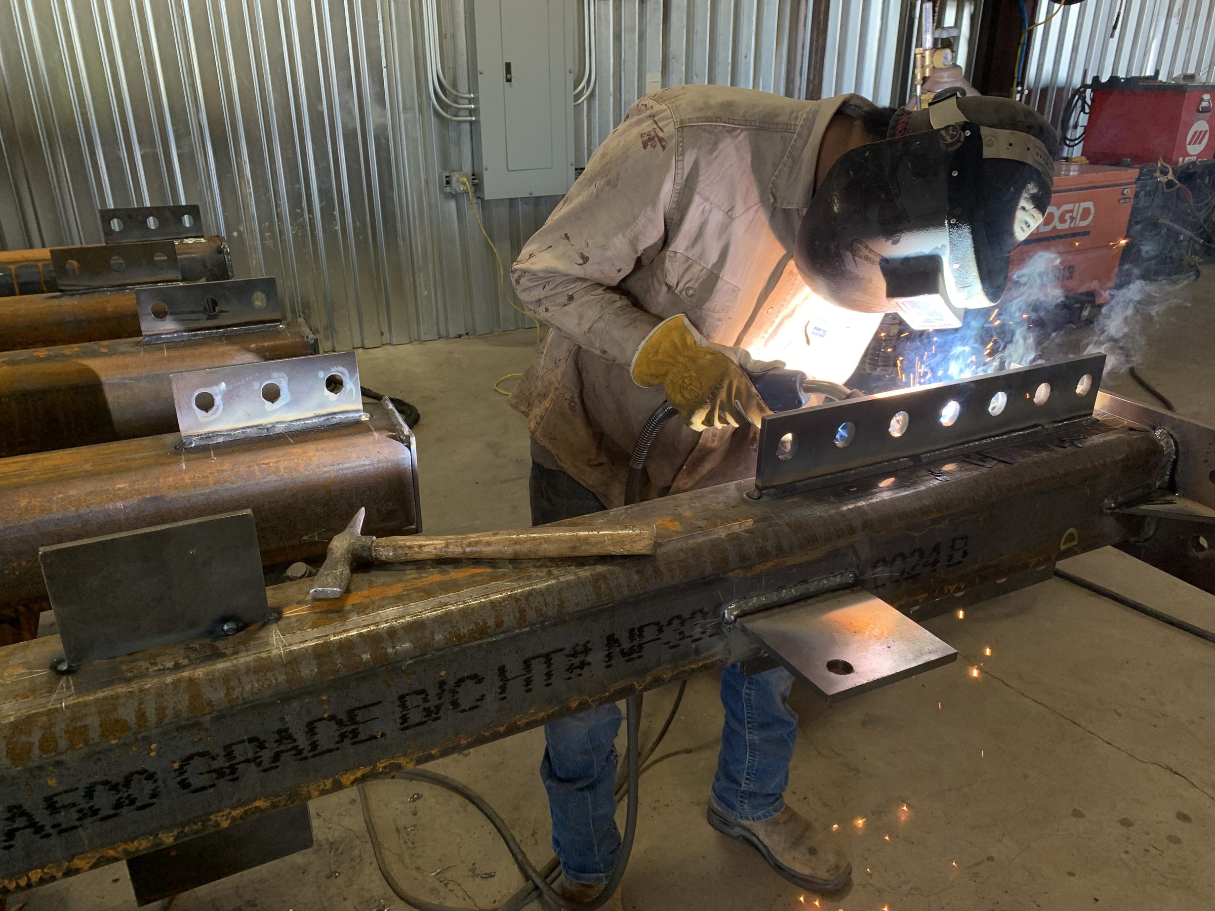 Welder attaching a bolted connection plate to an A500 steel tube column, with finished columns lined up beside him