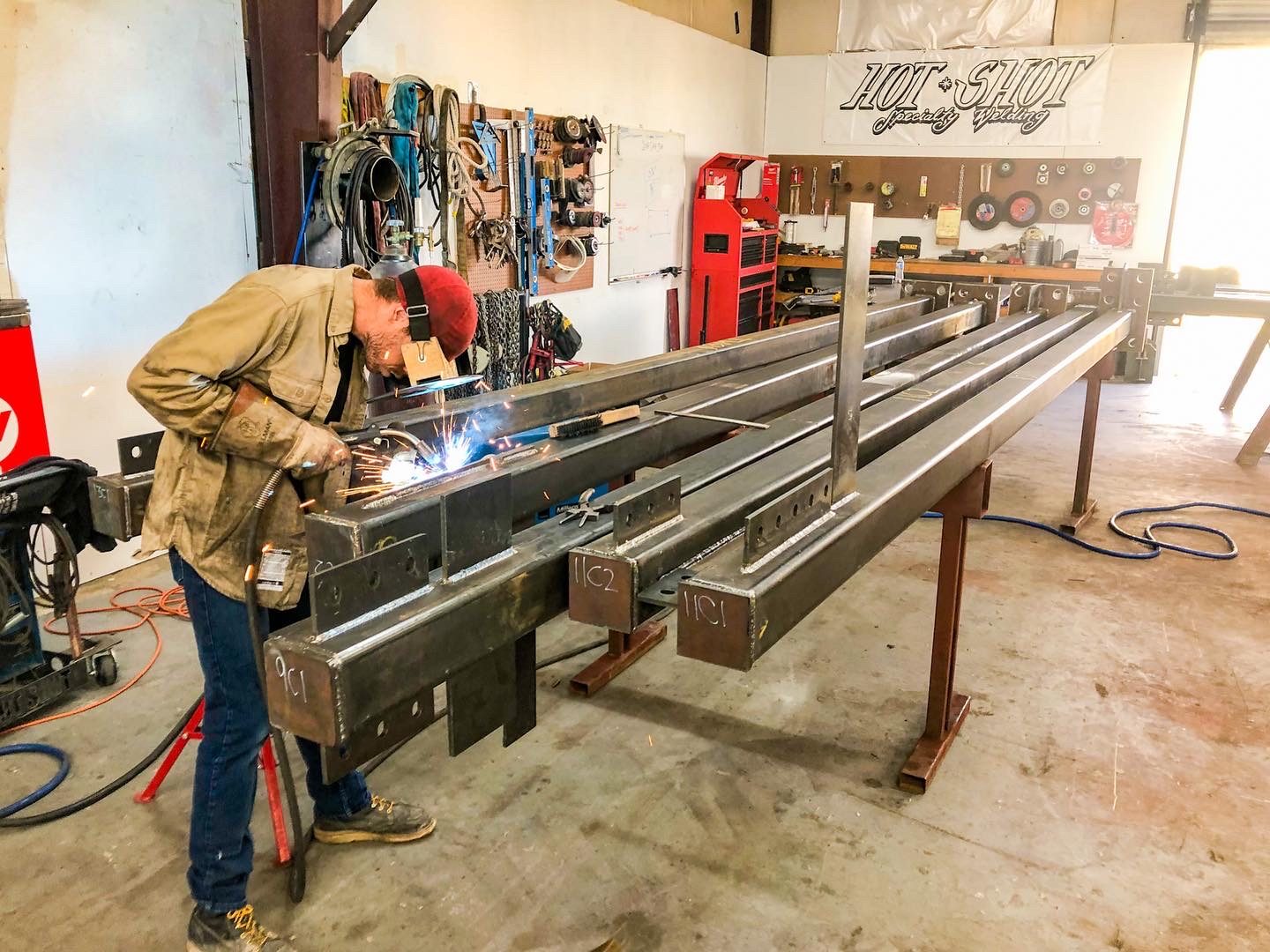 Welder running a bead on a set of labeled HSS steel columns on shop stands, with the Hot Shot banner on the back wall
