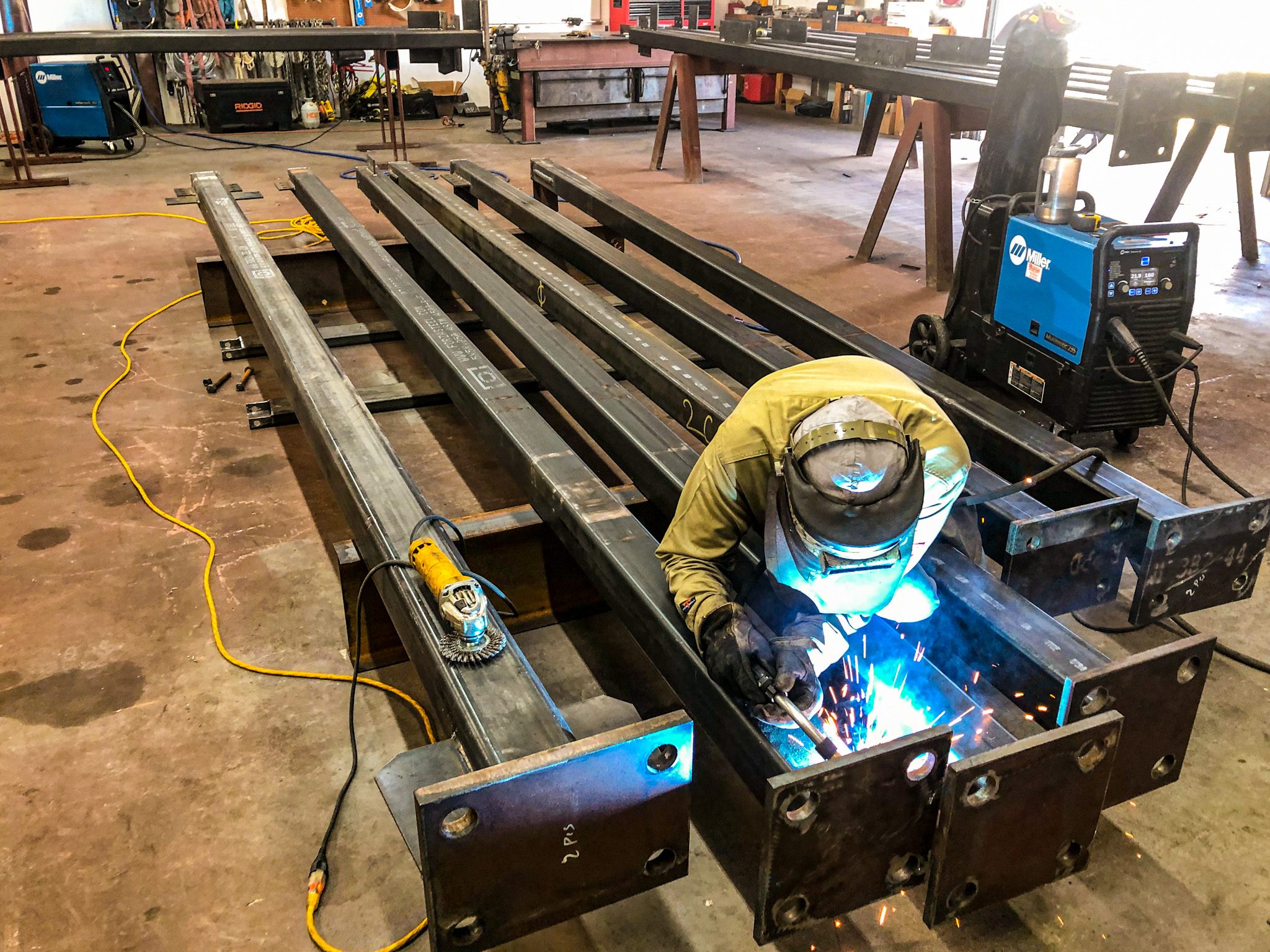 Welder fitting base plates to a row of rectangular steel tube columns on the shop floor, with a Miller welder beside him