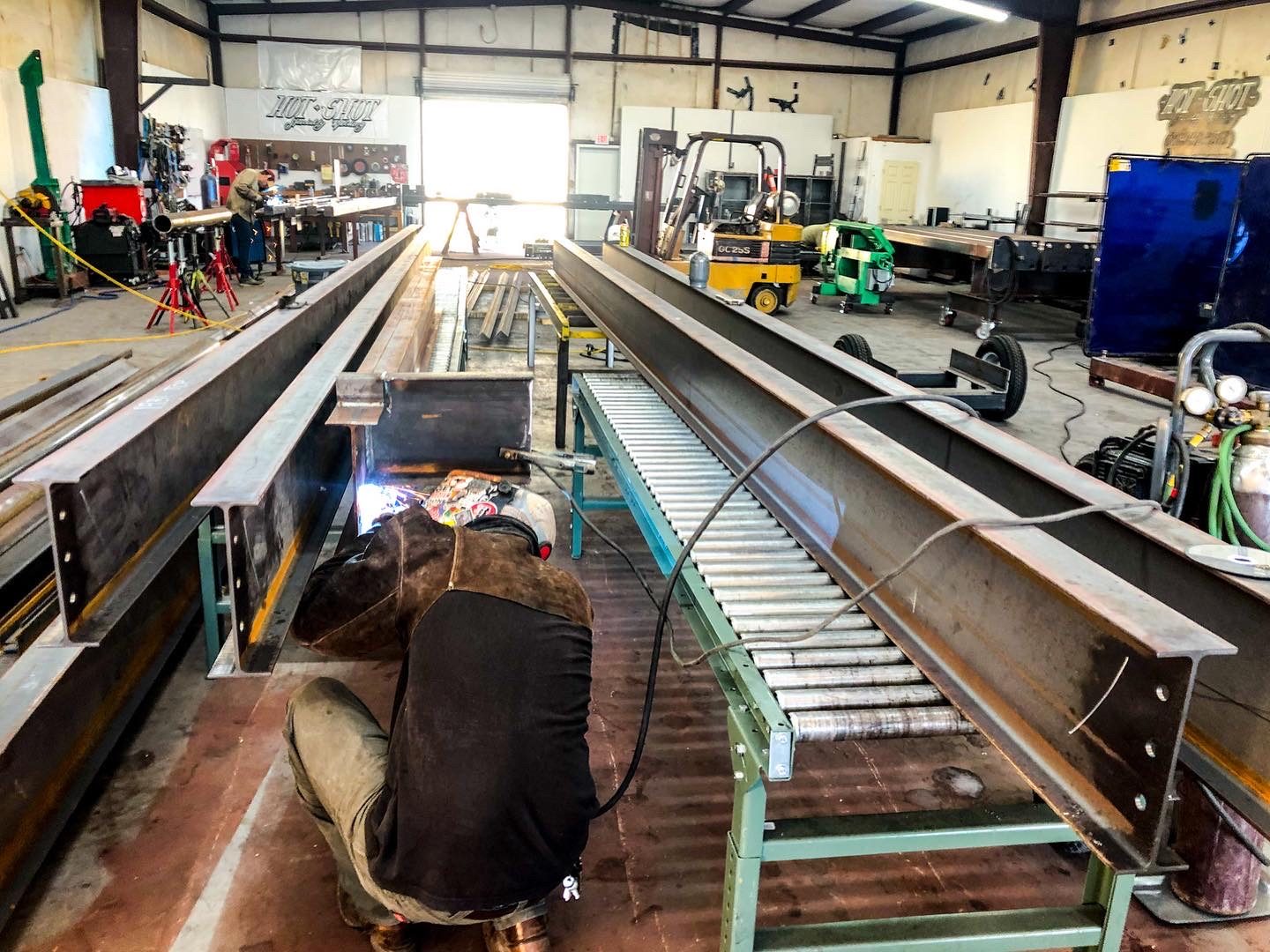 Welder working between wide-flange steel beams on a roller conveyor, with a forklift and the full fab shop behind him