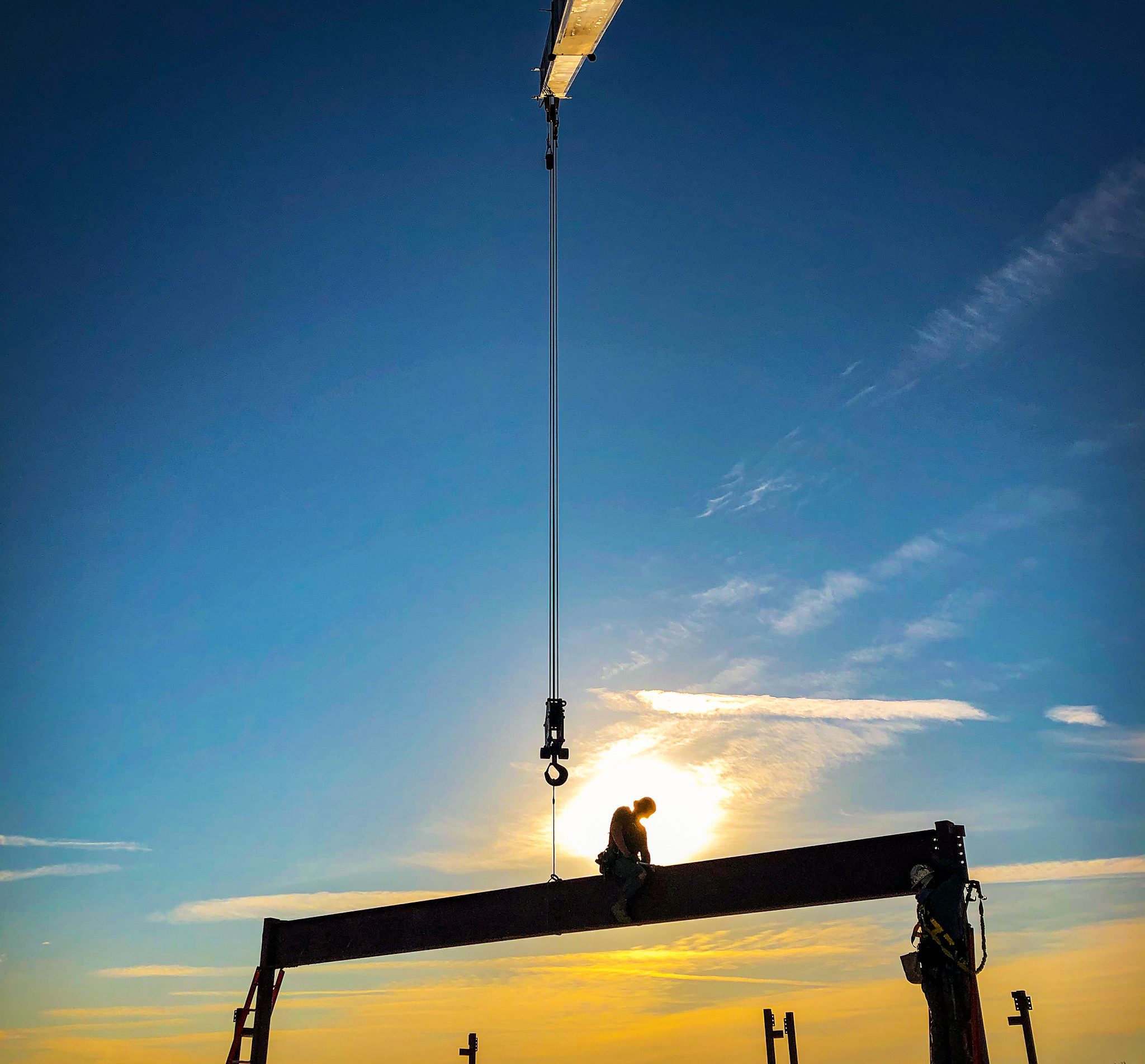 Ironworker seated on a steel beam hanging from the crane hook, silhouetted against a sunset sky